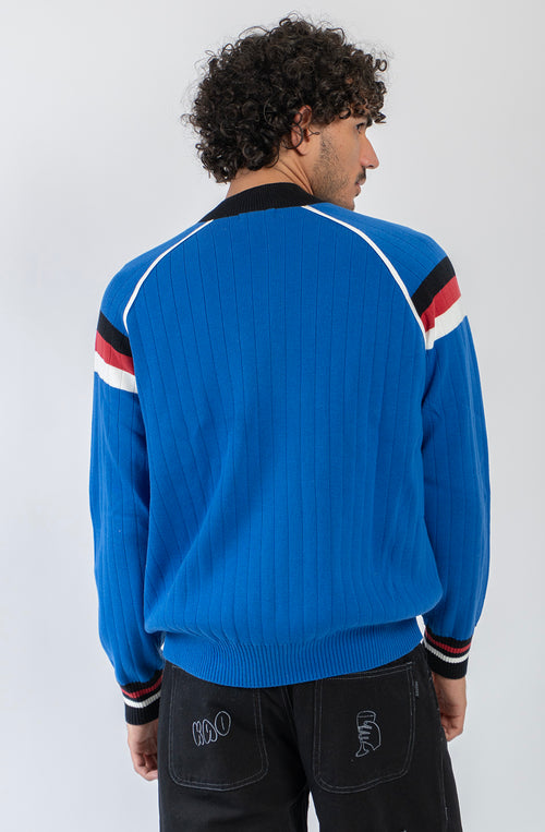 Chaqueta de punto azul con cremallera, cuello bomber y detalles deportivos en blanco, negro y rojo.