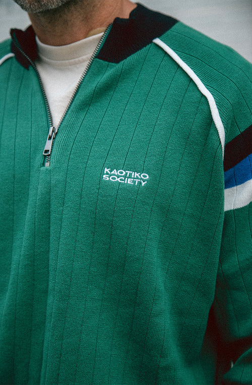 Jersey de punto verde con media cremallera, cuello alto y detalles deportivos en blanco, negro y azul.
